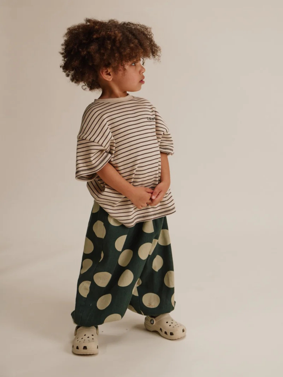A young child stands sideways wearing the EMERSON KALE POLKA TROUSER, featuring wide dark green pants with large beige polka dots, paired with a beige striped shirt and beige slip-on shoes against a plain background.