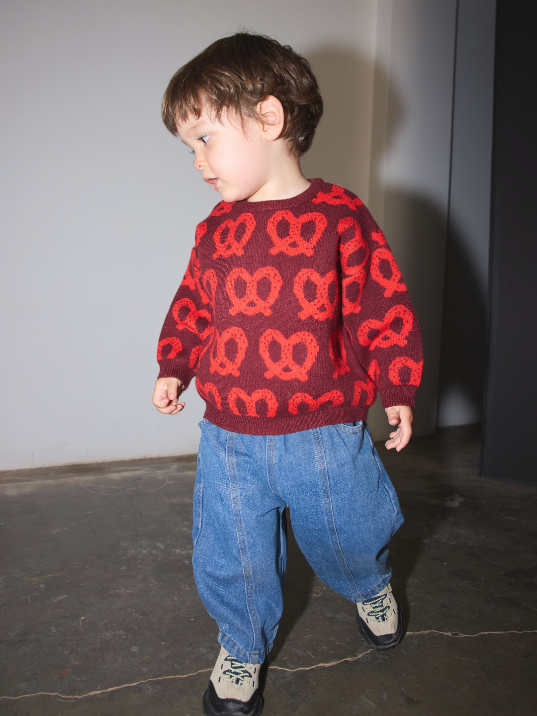 Red | A toddler wearing a red and dark red pretzel-knit sweater, loose blue jeans, and sneakers, standing in an indoor space.