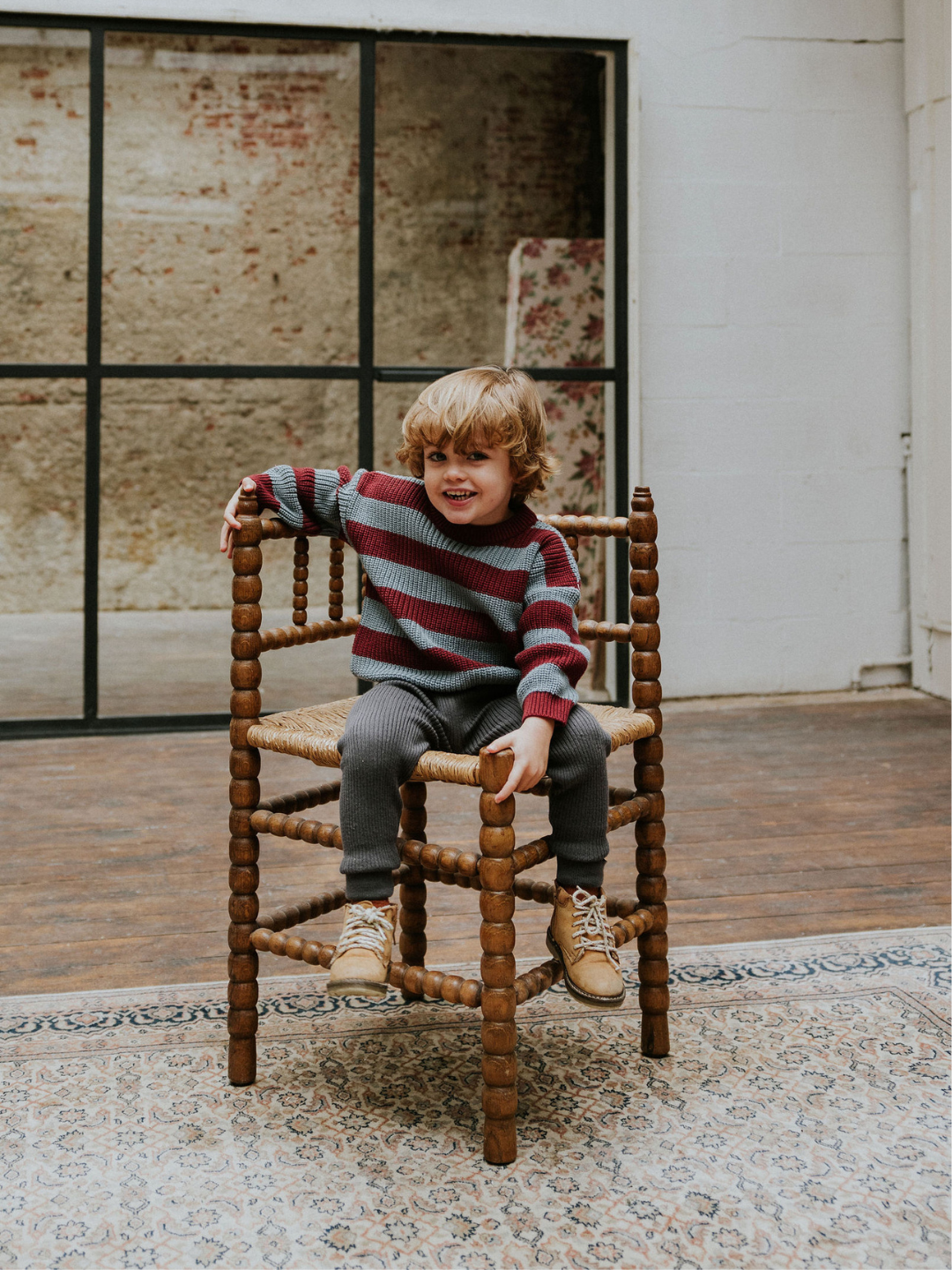 A small child with curly hair sits on a wooden chair with intricate carvings, smiling and dressed in the STRIPES SWEATER, gray pants, and brown shoes in an indoor space with a patterned rug.