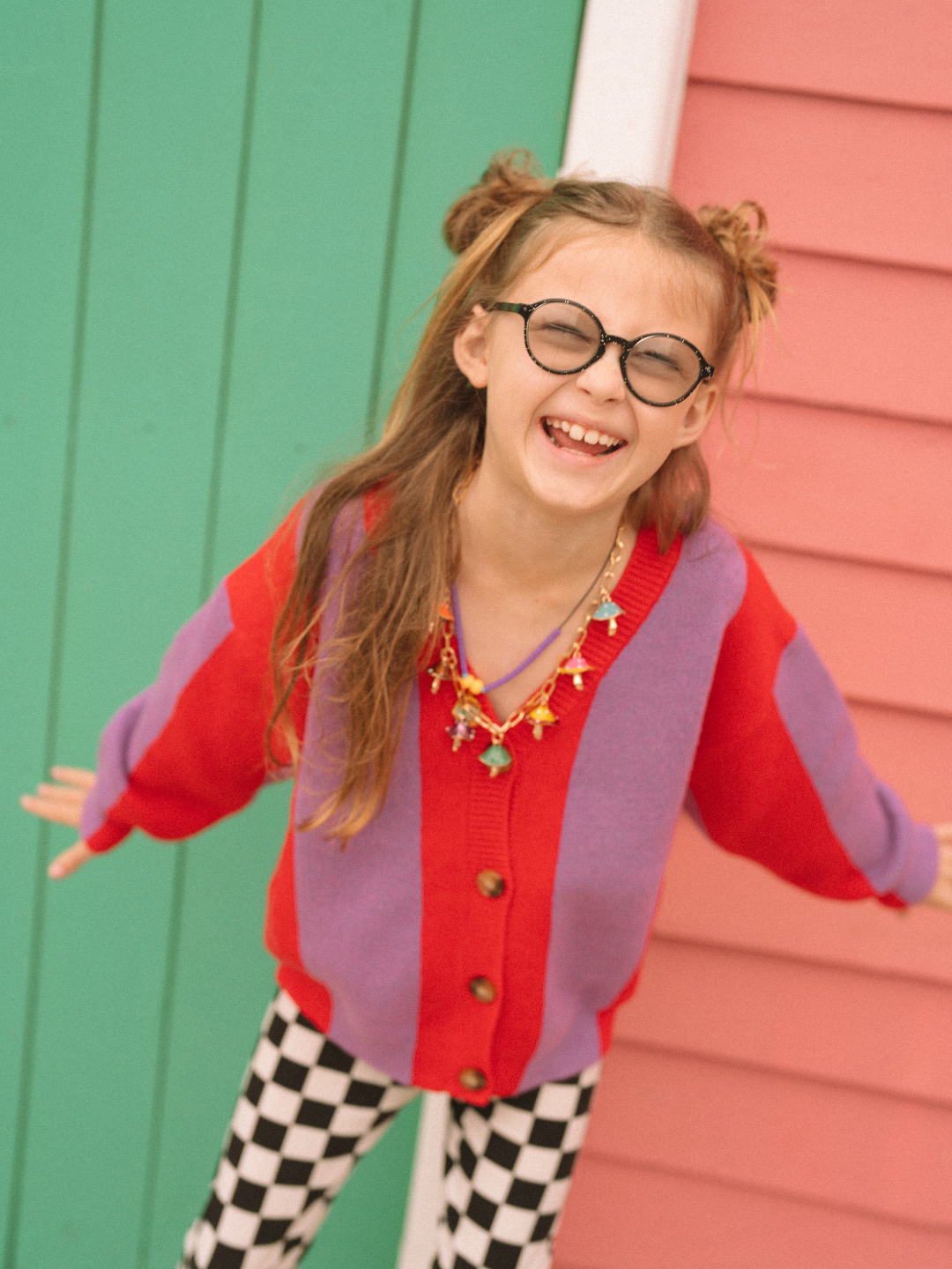 Red | Young girl wearing a Red Stripe cardigan and checkered pants standing in front of a green and pink wall.