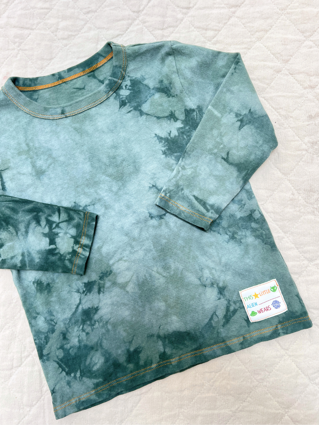 Sage | The COSMO TIE DYE LONG SLEEVE in green with yellow stitching lies flat on a quilted white surface, featuring a small fabric label at the bottom hem—crafted in Los Angeles.