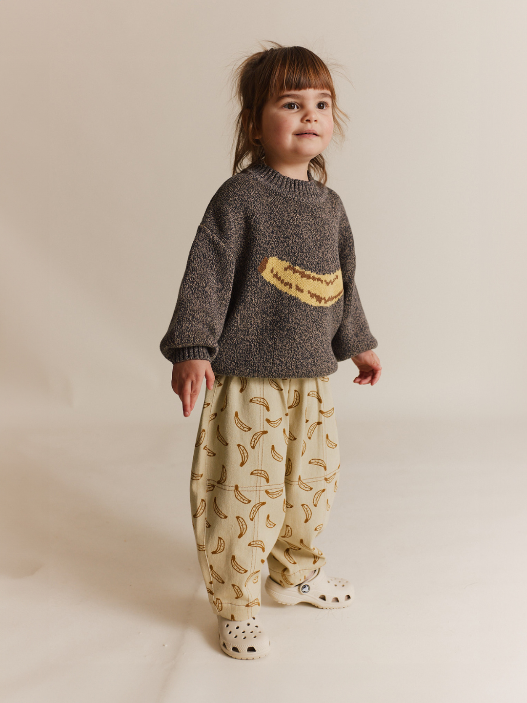 A young child stands on a plain background wearing a gray sweater with a yellow patch, RIAN BANANA TROUSER—oversized balloon leg trousers in banana-patterned organic cotton twill—and white slip-on shoes.