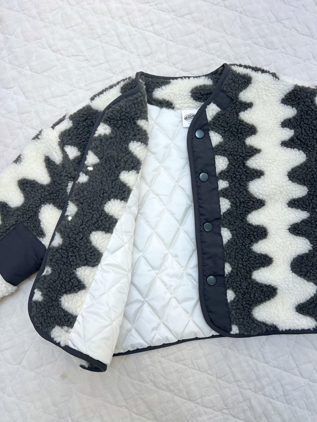 The WAVELENGTH SHERPA JACKET features a black and white zigzag pattern, snap buttons, and a cozy sherpa fleece exterior, laid open to display its white quilted interior on a light-colored quilted surface.