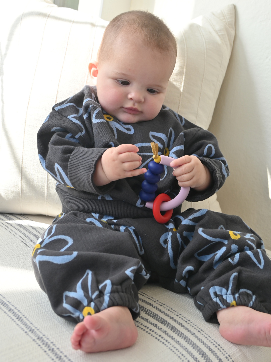 A baby sits on a striped cushion, wearing a dark floral outfit and looking at the colorful MT X JM ARCH RING TEETHER—this sensory-friendly teether made in the USA is ideal for little hands and curious minds.