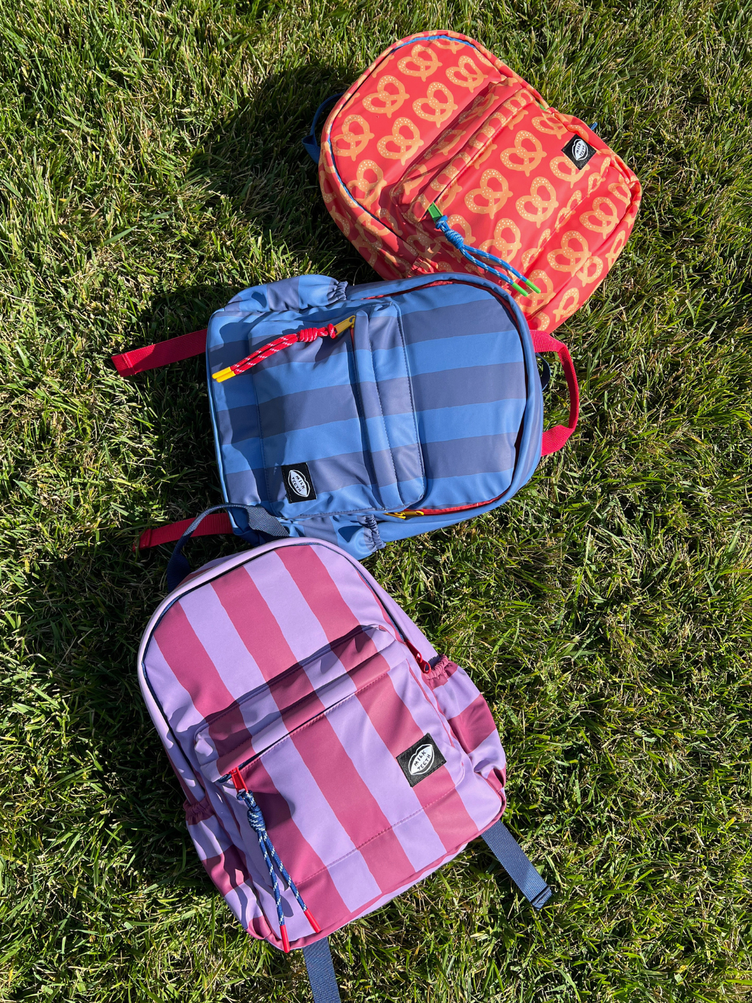 Purple Stripe | Three colorful kids backpacks from Milk Teeth are arranged diagonally on a green grass lawn. The top backpack is orange with a yellow pretzel pattern, the middle is blue with darker gray stripes, and the bottom is maroon and purple with vertical stripes.