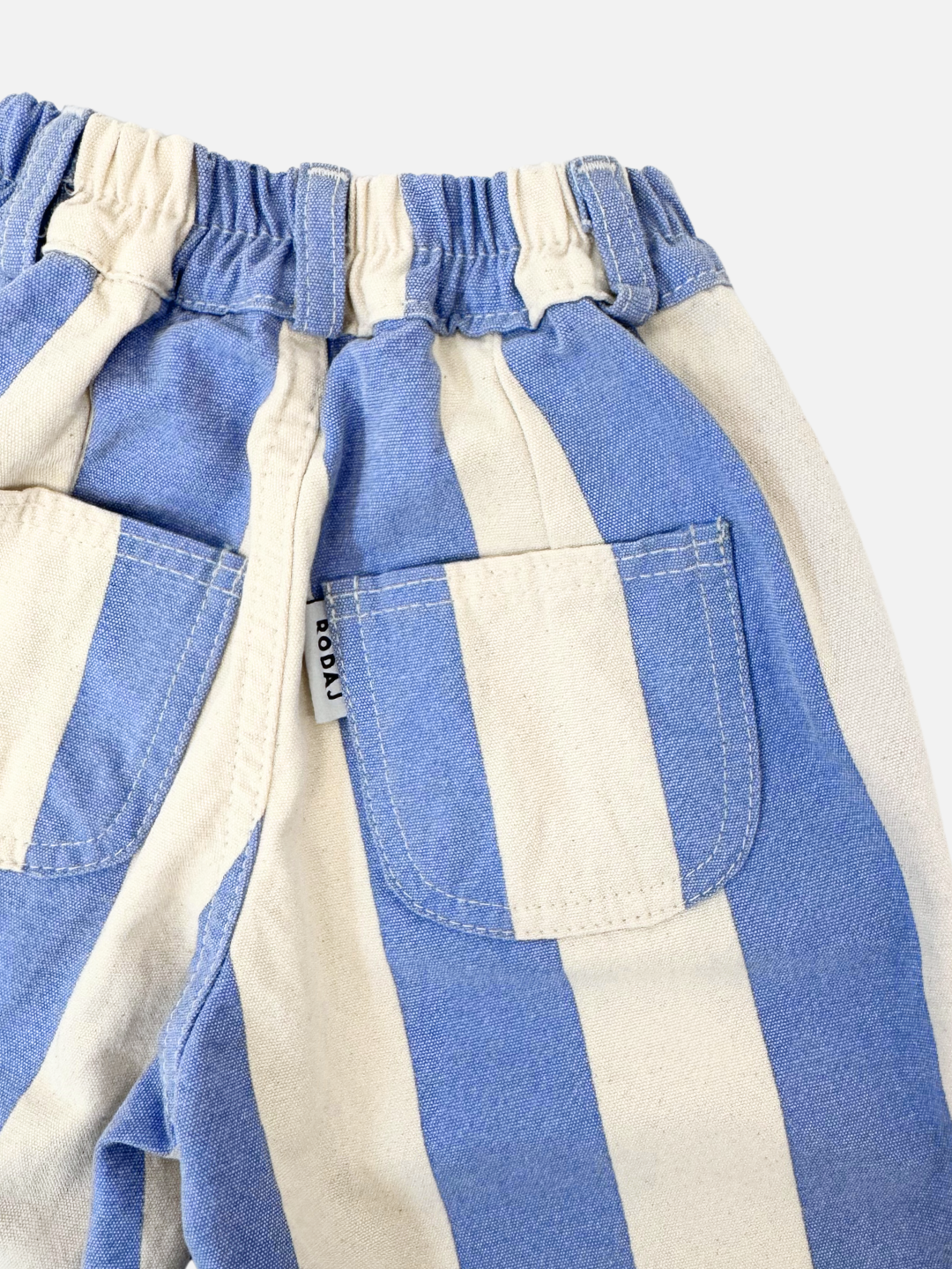 The CABANA STRIPE PANTS are blue and white vertically striped, high-waist with two back pockets and an elastic waistband. Made from soft cotton canvas as part of a Korean children's brand collection, they feature a small tag with text by one pocket.