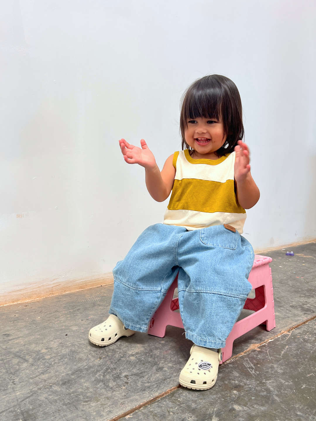 Cream/Gold | A young child smiles and claps hands while wearing the TRACK TANK, a vintage sportswear-inspired yellow and white striped top, paired with blue jeans, sitting on a small pink stool.