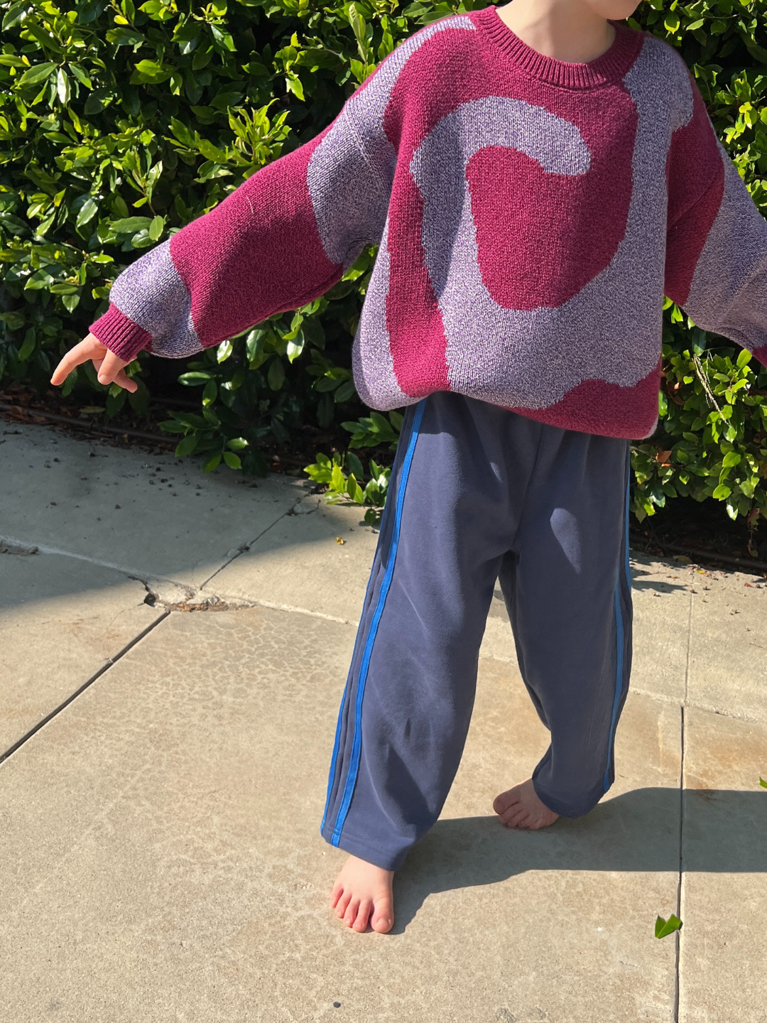 Navy | A child stands barefoot on a sunlit sidewalk, wearing SUNDAY TRACKPANTS with side stripes and a red patterned sweater, lush greenery in the background.