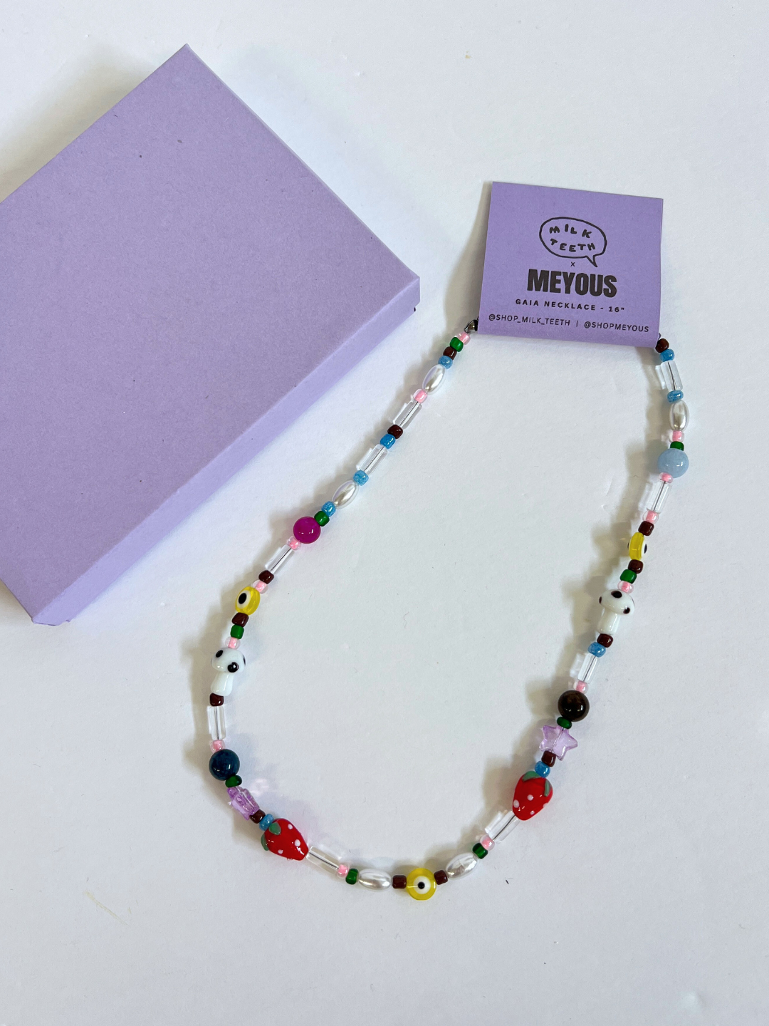 The GAIA NECKLACE, featuring vintage glass beads plus handmade strawberry and mushroom beads, is displayed beside a closed lavender box and a matching "MEYOUS" product tag.