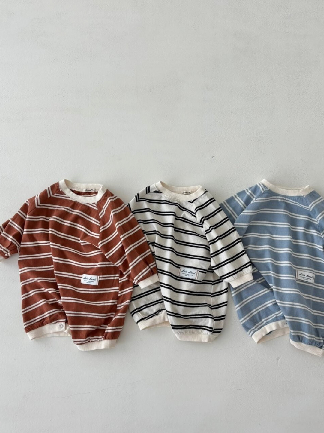 Three SLOUCHY STRIPE ROMPERs by a Korean baby brand, made of cotton jersey with white stripes, are laid side by side in brown, white, and blue on a light background.