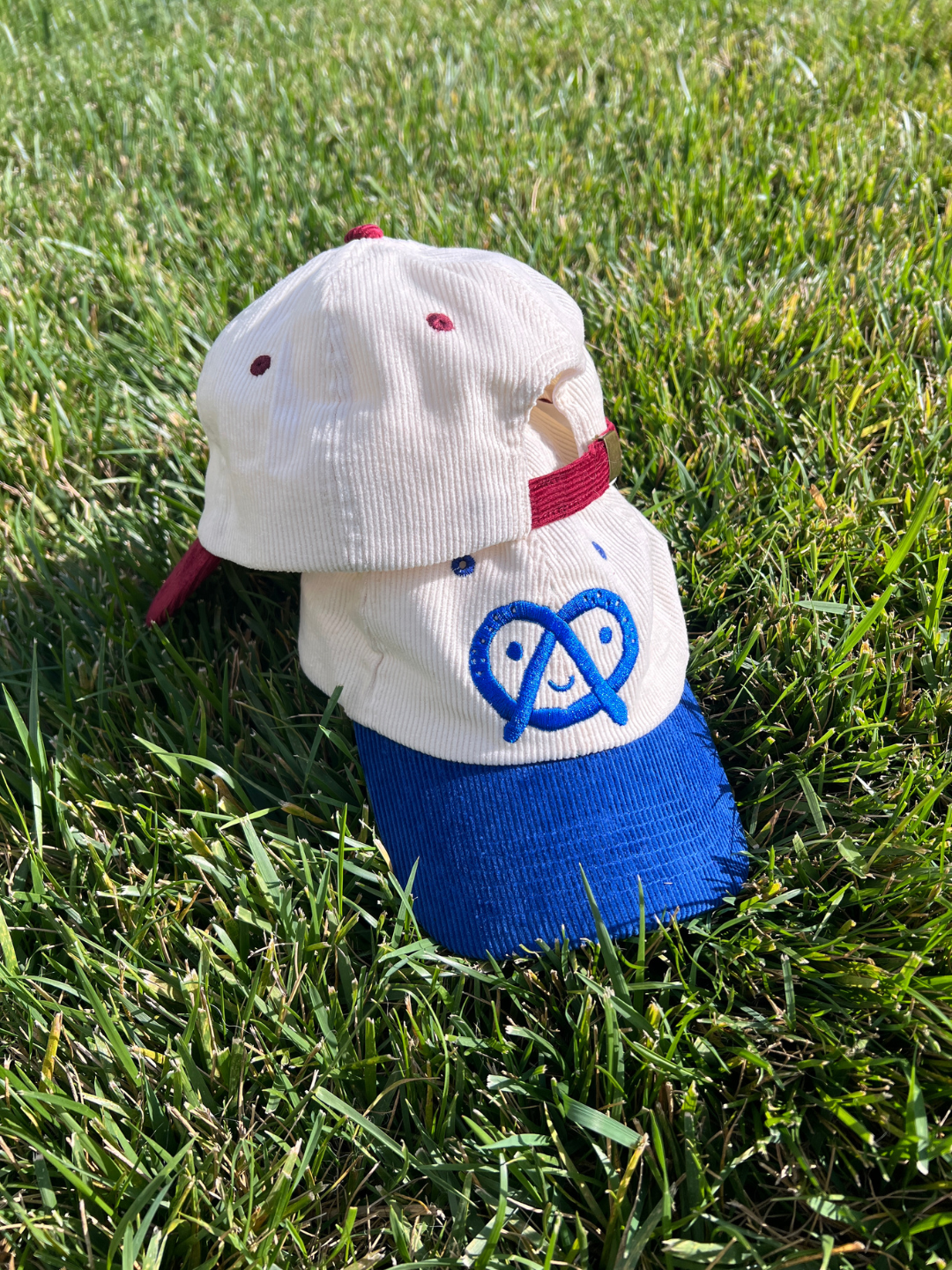 A white corduroy baseball cap with a blue brim and a blue embroidered pretzel logo, resting on green grass outdoors.