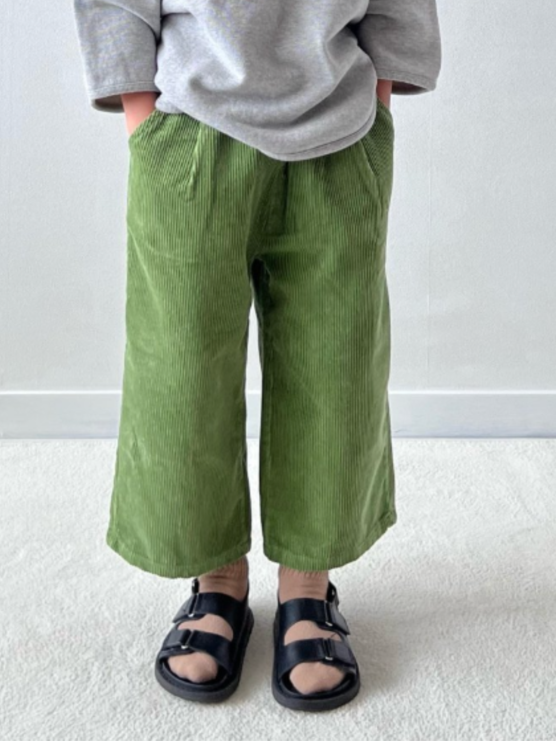 A waist-down cropped image of a child wearing wide-leg corduroy pants in moss green, worn with a grey sweatshirt, beige socks, and black sandals. The child is standing on grey carpet in front of a white wall, with hands in pockets.