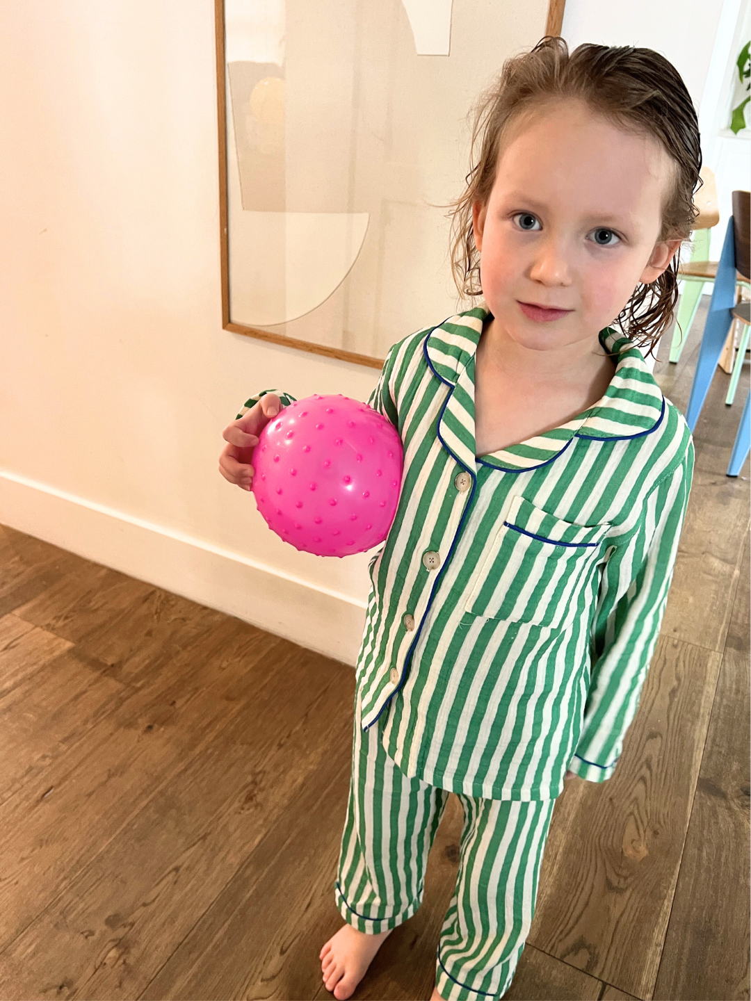 Green Stripe | A child wearing the MOVIE NIGHT SET, a green-striped cotton muslin set, holds a pink spiky ball while standing on an indoor wooden floor.