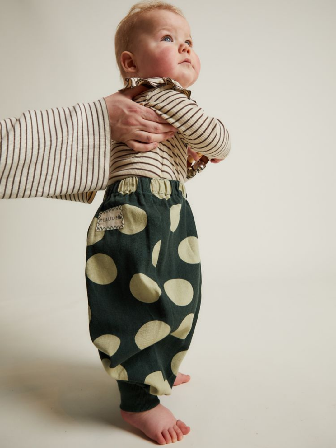 An adult holds a baby dressed in a striped shirt and EMERSON KALE POLKA TROUSER, featuring loose dark green pants with large light-colored polka dots, standing on a plain background.