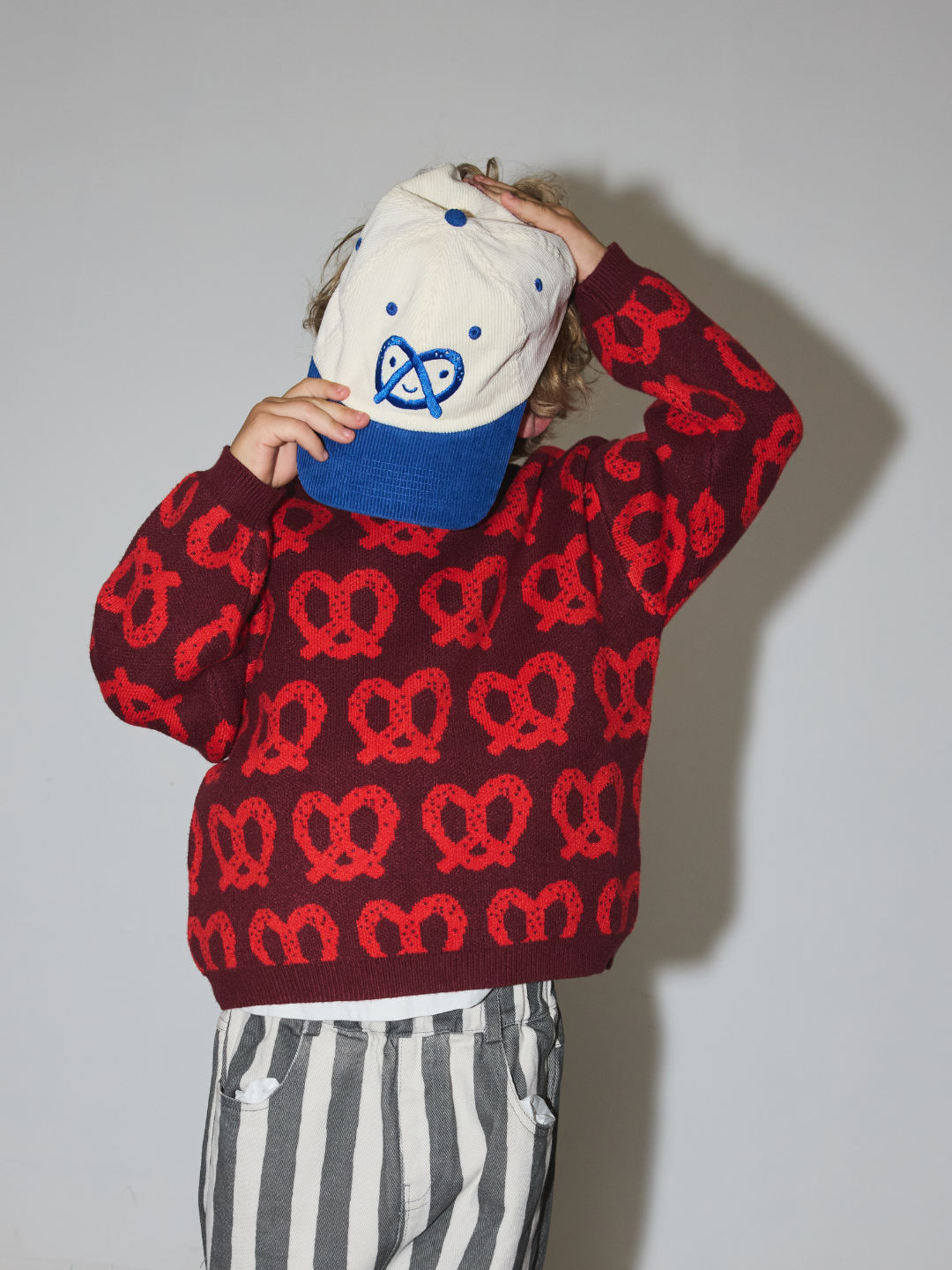 Red | A child wearing a burgundy sweater with a red pretzel pattern, striped pants, and a cream and blue baseball cap, posed against a plain gray wall.