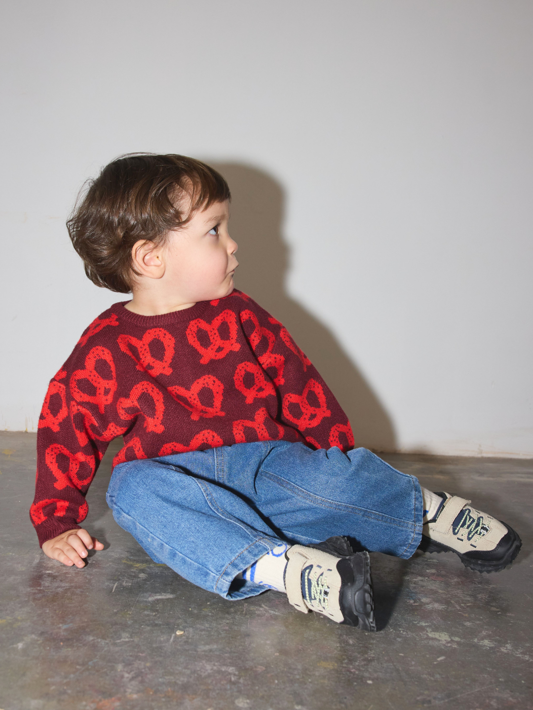 Red | A toddler sitting on a concrete floor against a plain light gray wall, wearing a burgundy knit sweater with a red pretzel pattern, blue jeans, and white sneakers.