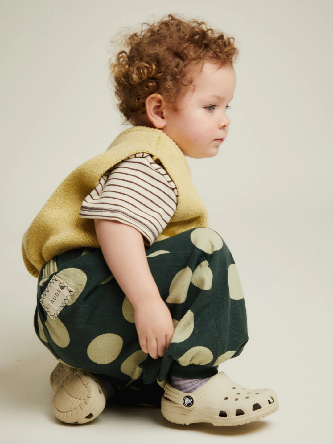 Toddler with curly hair squatting, wearing a yellow vest, striped shirt, EMERSON KALE POLKA TROUSER, and cream-colored clogs.