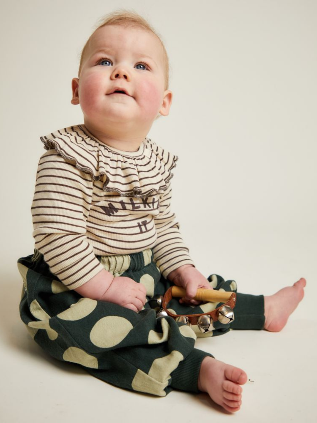 A baby sits on the floor wearing a striped shirt and EMERSON KALE POLKA TROUSER, holding a tambourine and looking upward against a plain background.