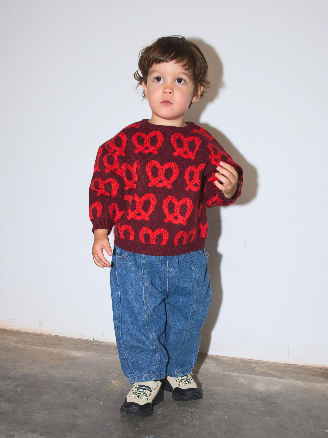 A toddler standing against a plain white wall wearing a dark red knit sweater with a red pretzel pattern, loose blue jeans, and light-colored sneakers.