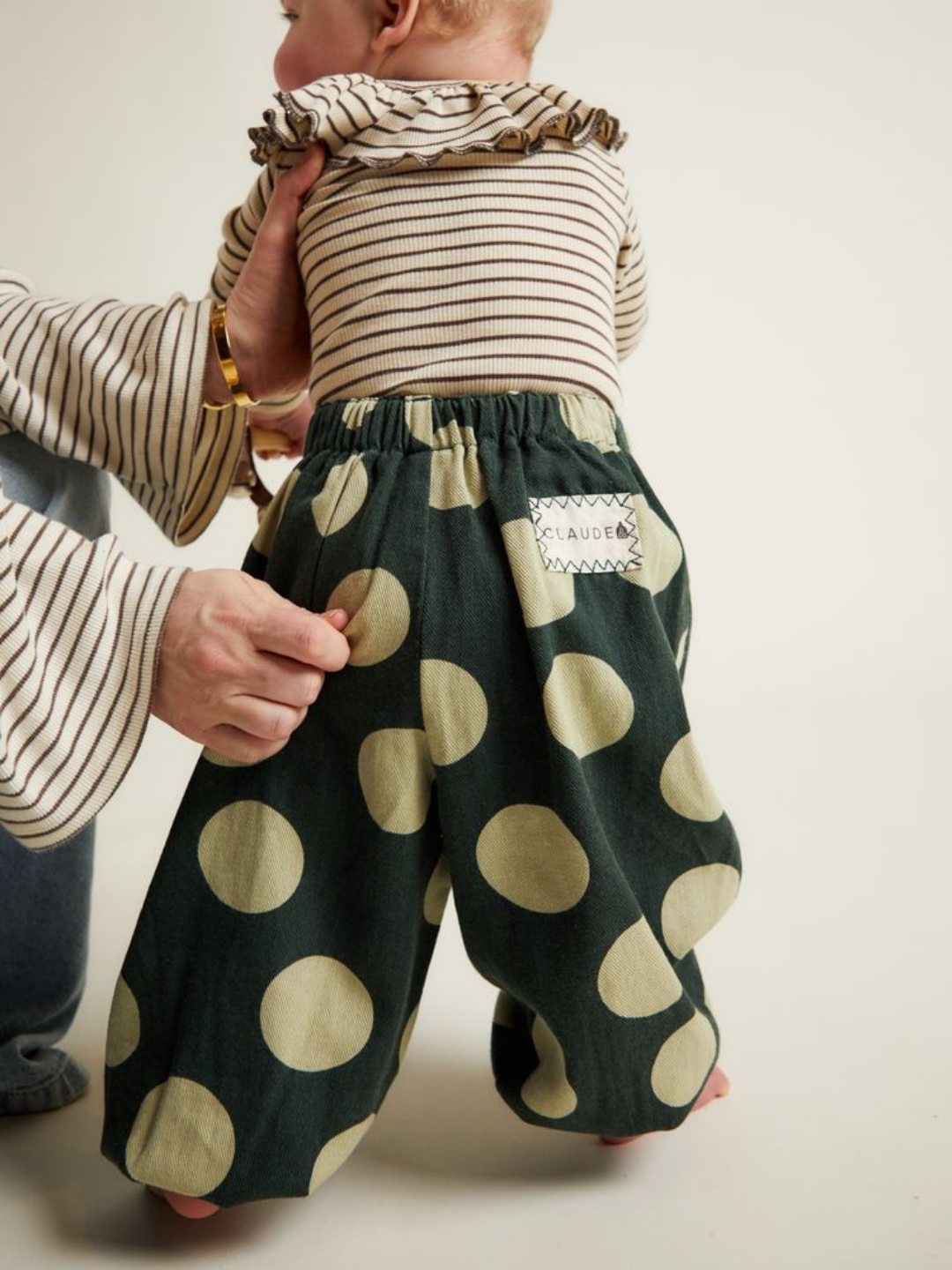 An adult helps a baby labeled “CLAUDE,” dressed in a striped top and EMERSON KALE POLKA TROUSER—green pants with large beige polka dots—to stand.
