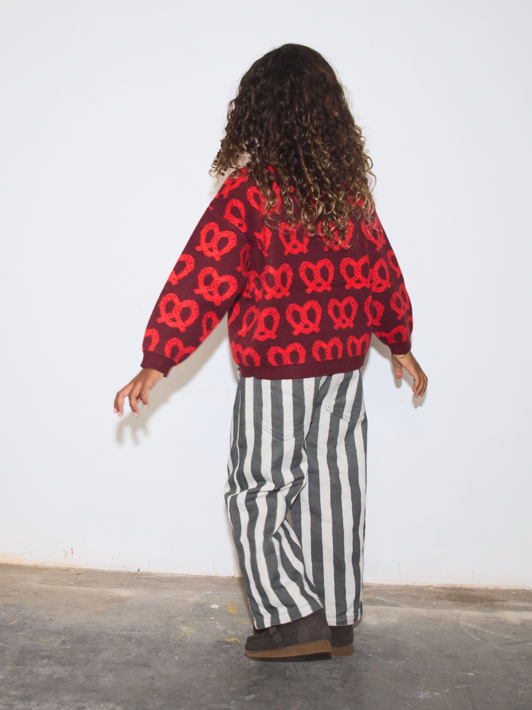 A child shown from the back wearing a red knit sweater with a red pretzel pattern, paired with black and white vertically striped pants and brown shoes, standing against a plain white wall.