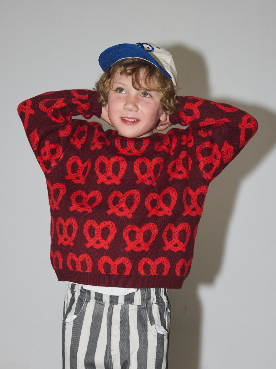 A young boy wearing a dark red knit sweater with a repeating red pretzel pattern, a blue and white baseball cap, and gray and white striped pants, posing with his hands behind his head against a plain light gray wall.
