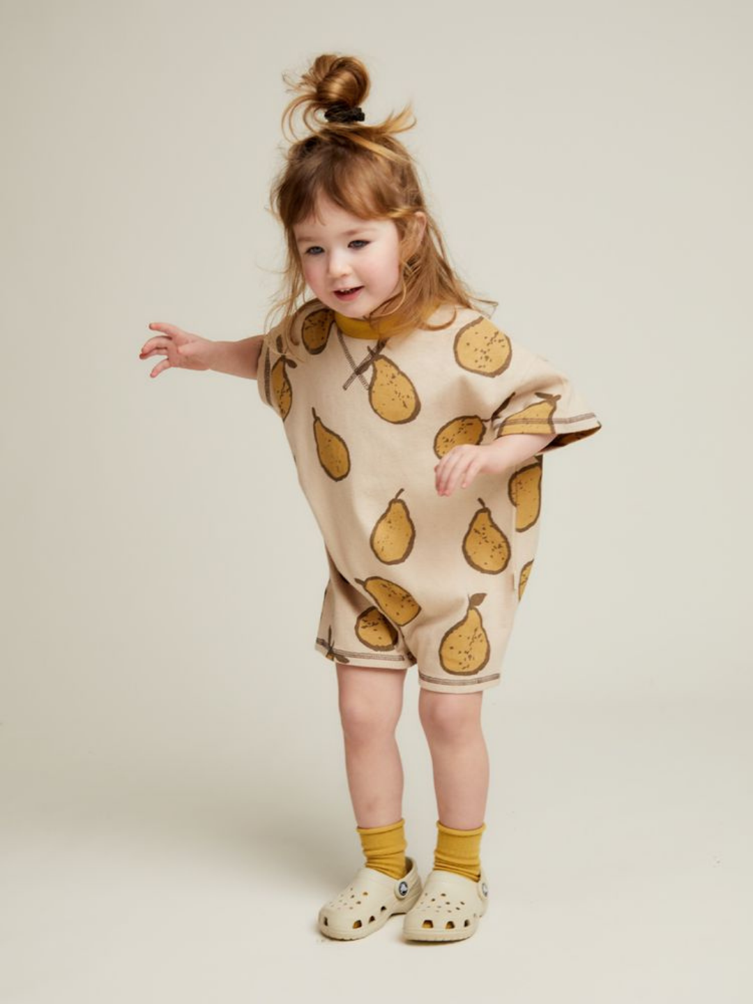 A young child smiles while wearing the OPAL ALMOND PEAR ROMPER—a loose beige outfit with a pear print—paired with yellow socks and light-colored clogs, standing against a plain backdrop.