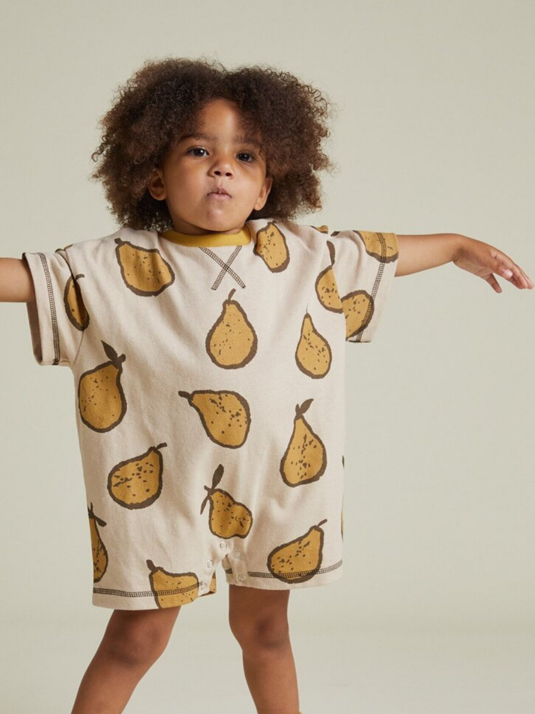 A young child stands with arms outstretched, wearing the OPAL ALMOND PEAR ROMPER— a beige romper adorned with yellow pear illustrations—set against a neutral background.