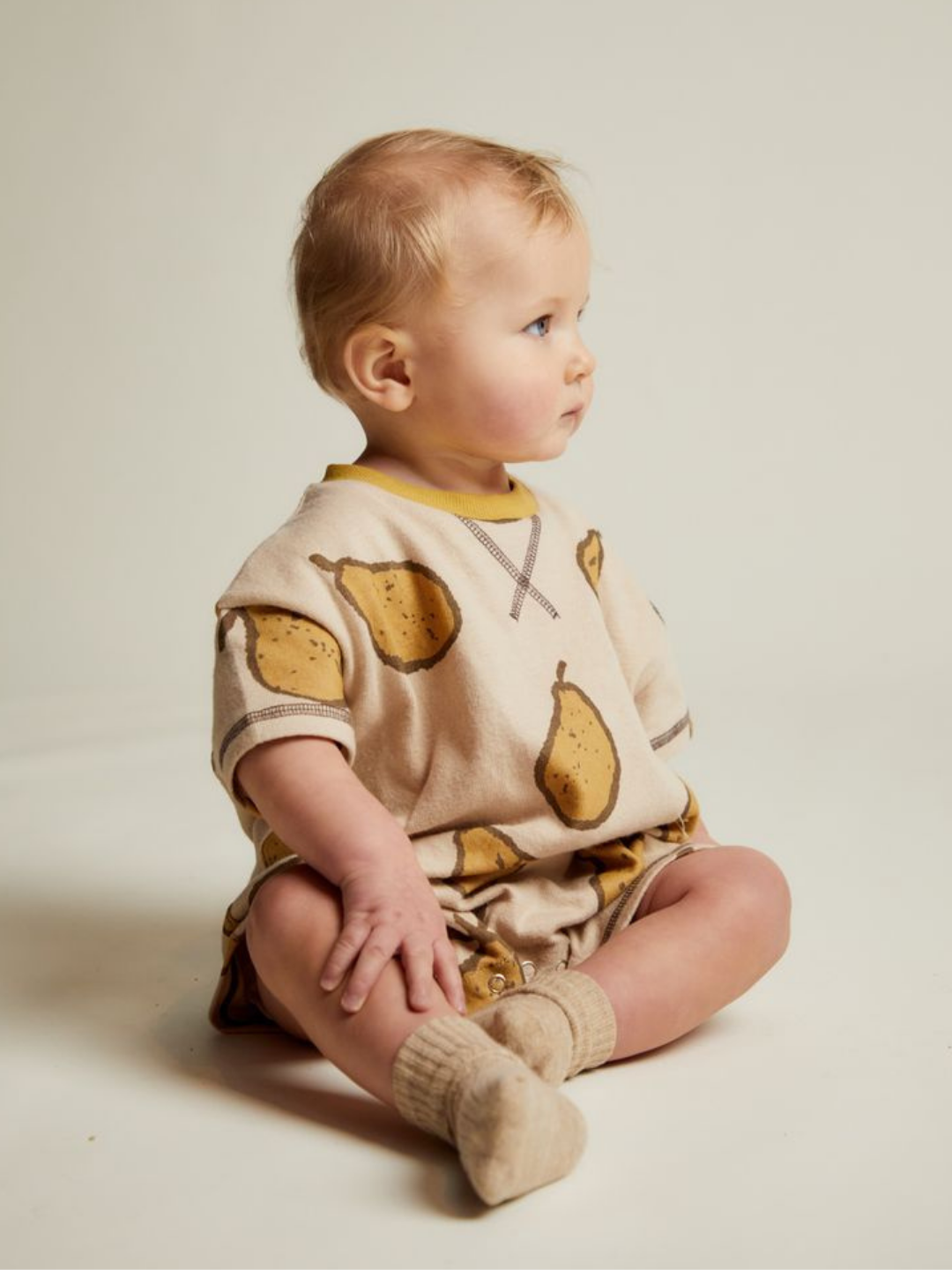 A baby sits on the floor in the OPAL ALMOND PEAR ROMPER with a pear print, beige socks, and looks to the side against a plain background.