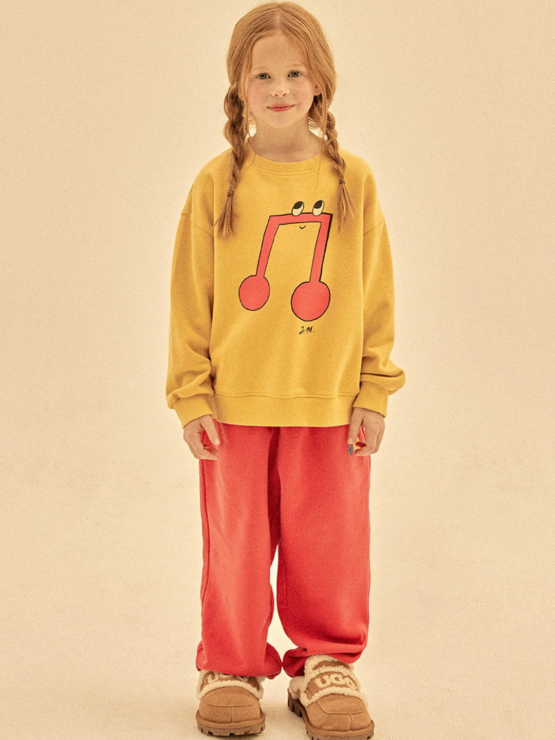 A young girl with braided red hair models the MELODY SWEATSHIRT, featuring a musical note graphic. She pairs it with red pants and beige shoes