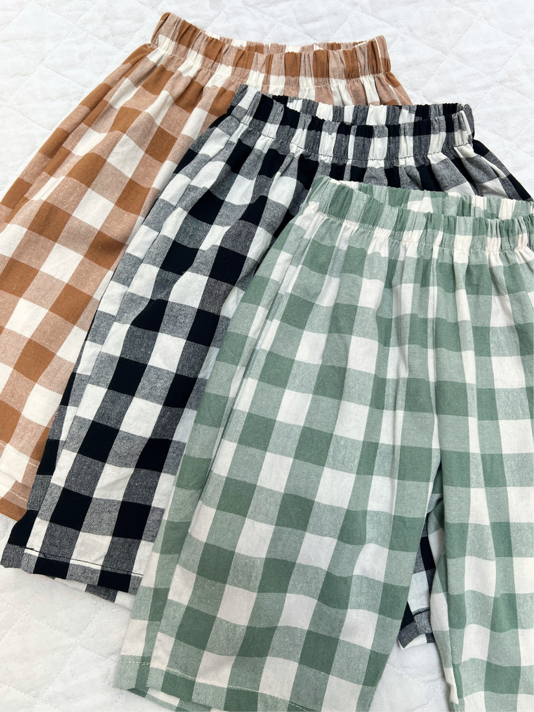 Three GINGHAM BABY PANTS with elastic waists in brown, black, and green are displayed on a white quilted surface—ideal for cozy Korean baby fashion.