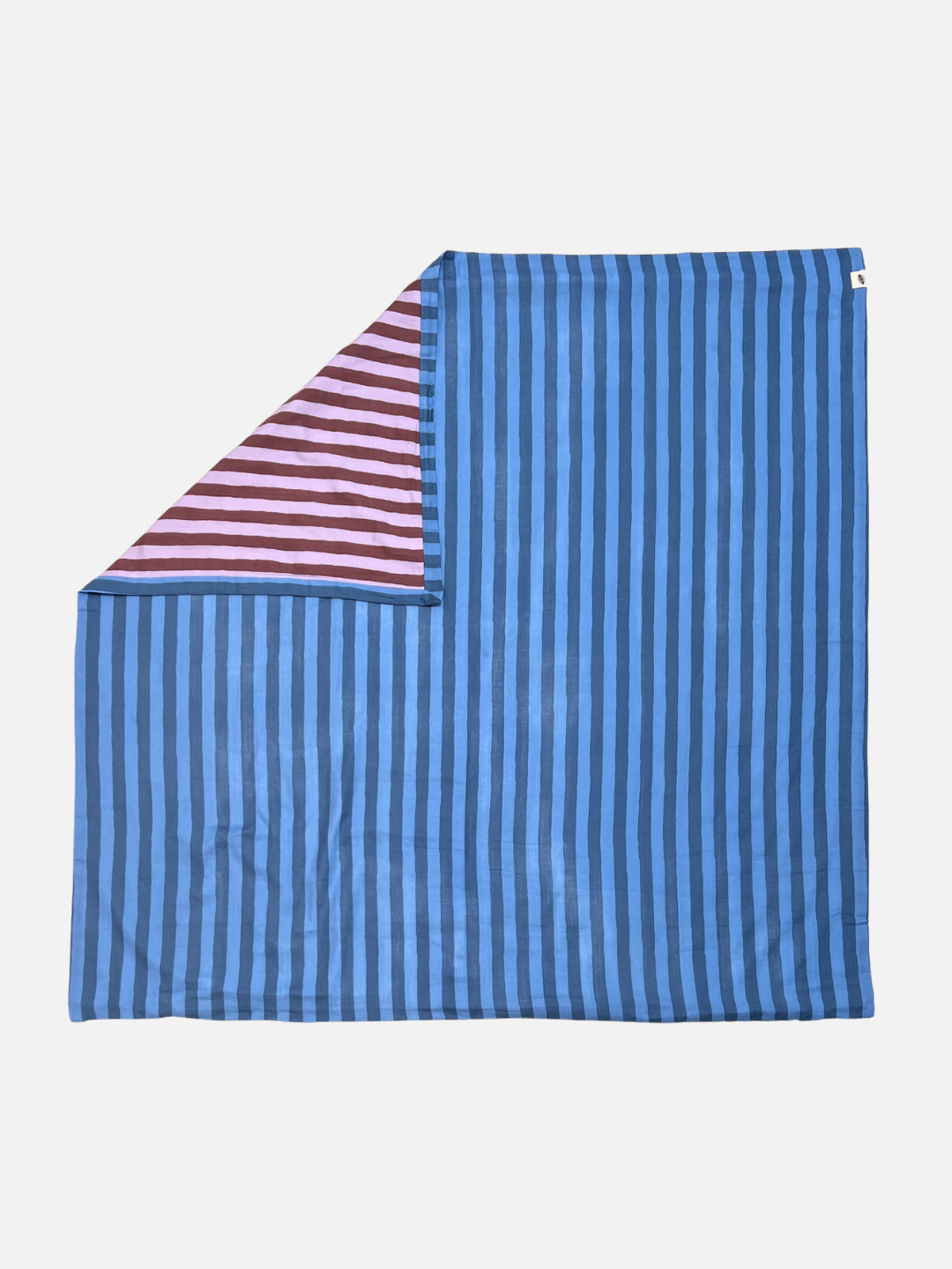 Blue Stripe | The organic cotton reversible quilt has medium blue and darker blue stripes, and is is shown flat on a light grey background, with a corner folded back revealing its purple striped underside.