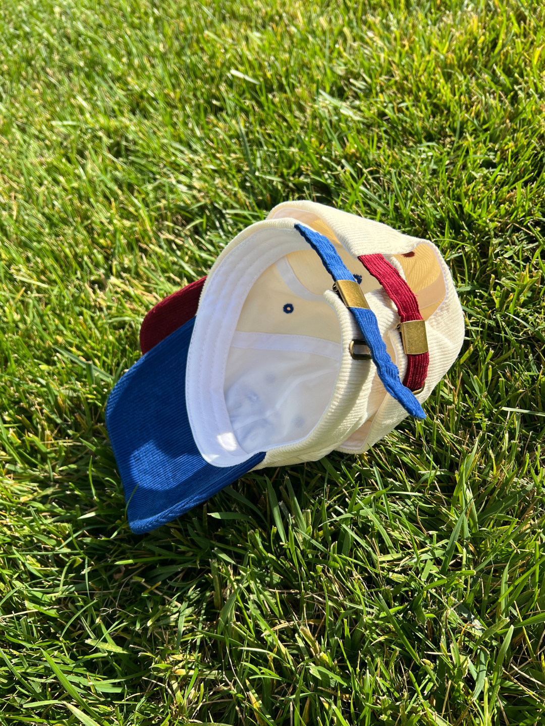 Two vintage-style two-tone baseball caps resting on green grass outdoors. The caps feature a cream-colored crown, a blue visor, and red and blue adjustable straps.