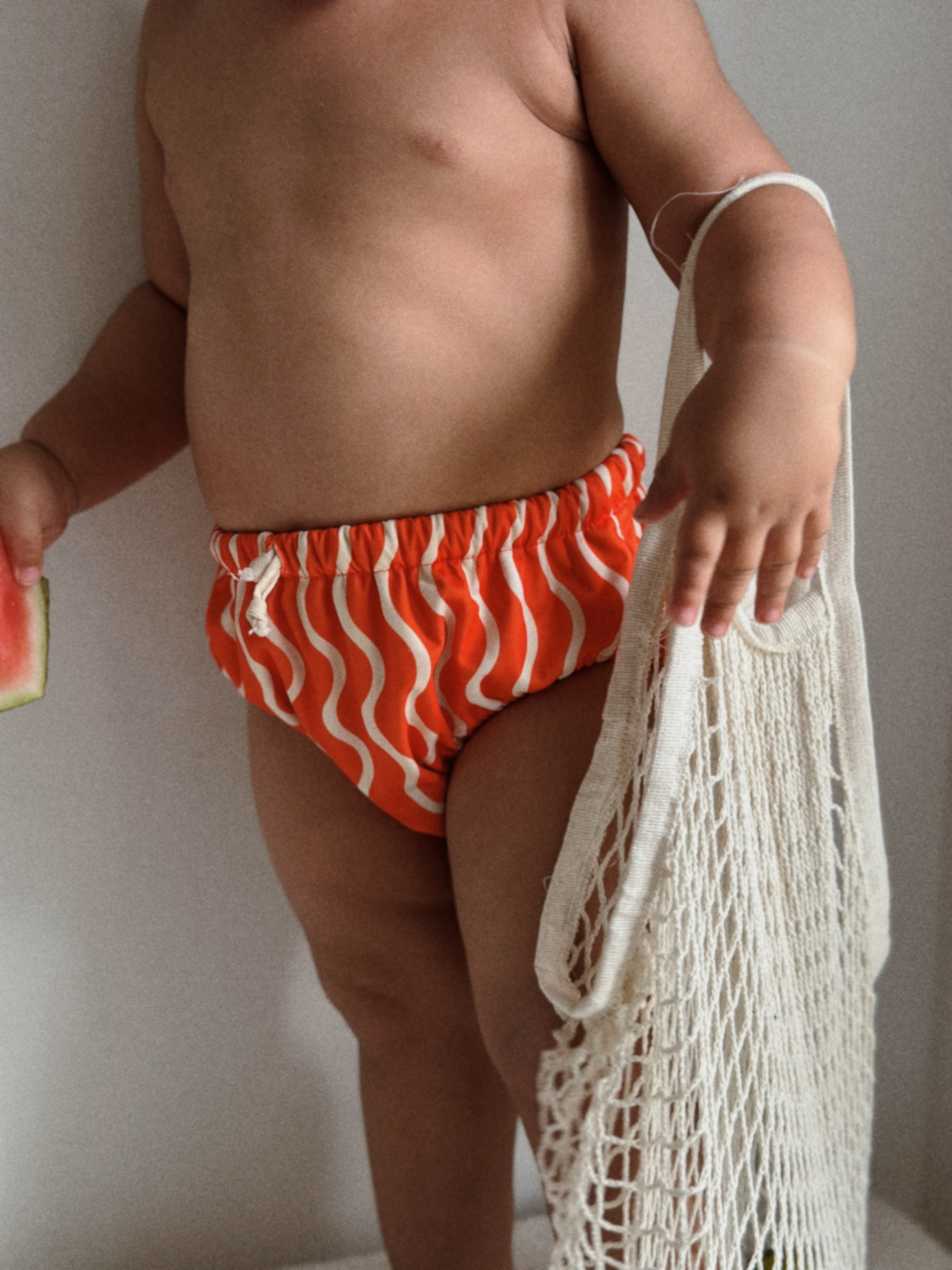 The front view of the swim diaper with an elastic waist with a tie and elastic leg holes on a child. The diaper is a bright orange with wavy and vertical cream colored lines.