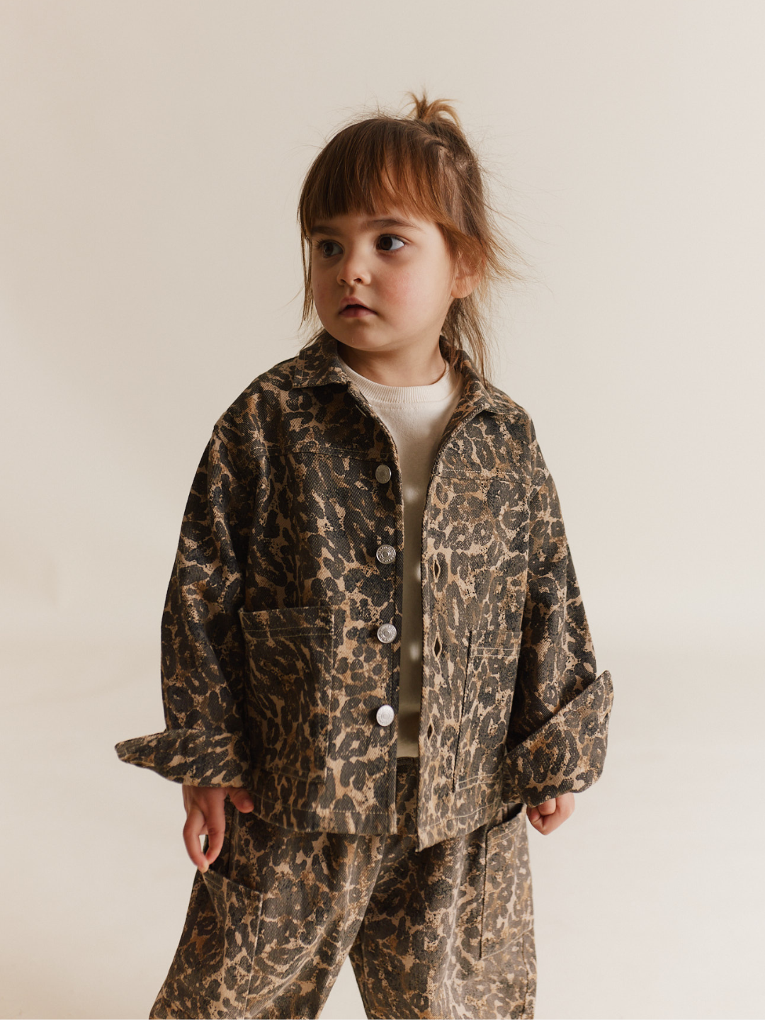 A young child in a FEN LEOPARD JACKET and matching pants made from organic cotton glances sideways with a calm expression.