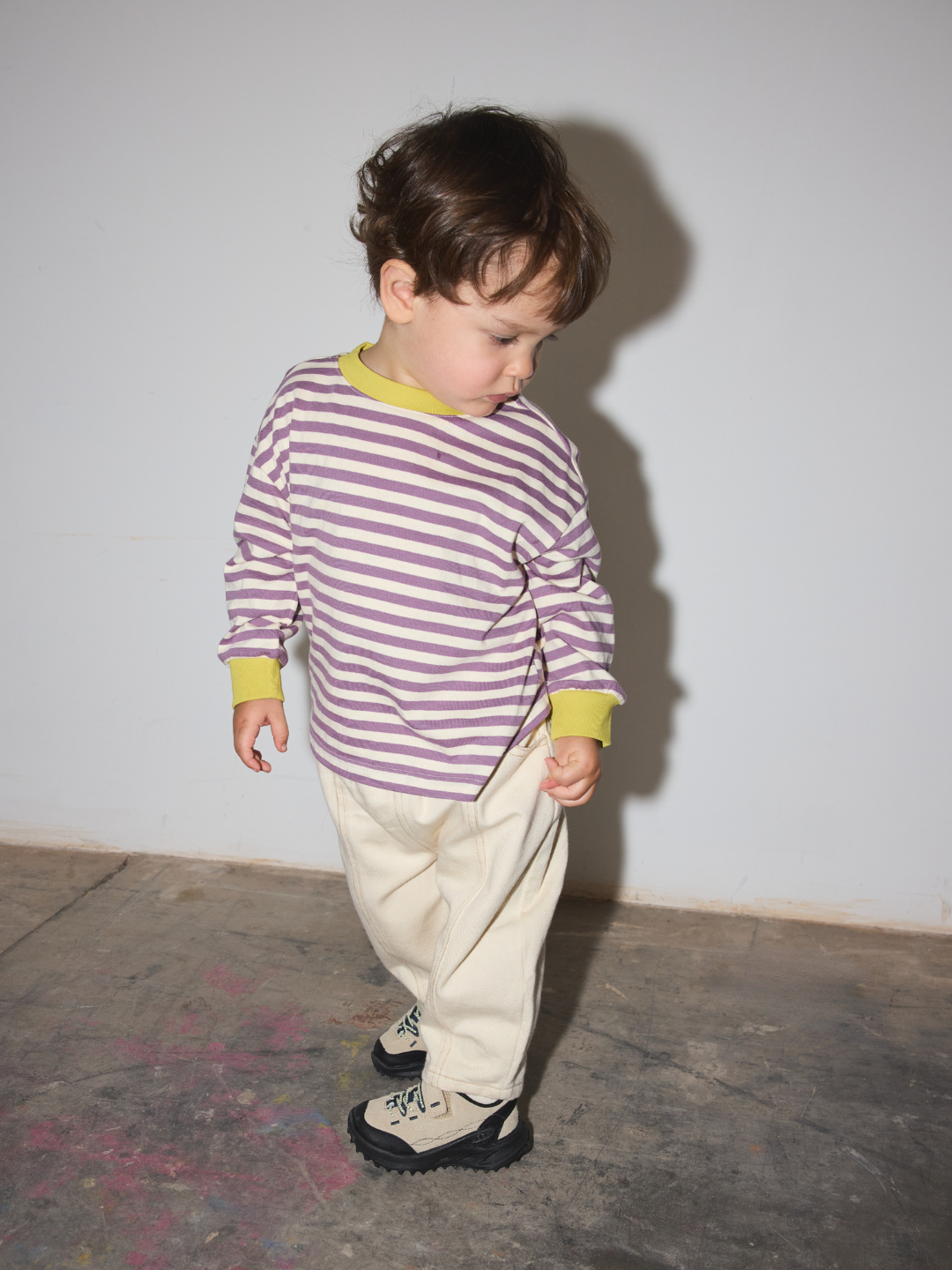 A toddler standing against a plain light gray wall, wearing a purple and white striped long-sleeve shirt with yellow cuffs, cream pants, and black-and-white sneakers.
