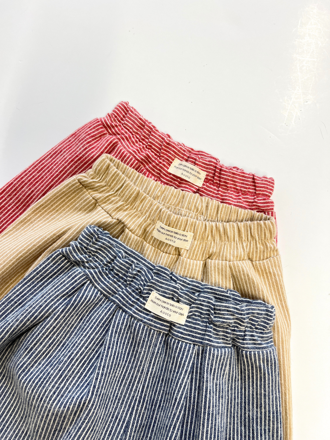 Navy | Three SOFT STRIPE BABY PANT pairs in red, beige, and blue with elastic waists and striped patterns are arranged in a row on a white background. Each features a sewn label at the waistband.