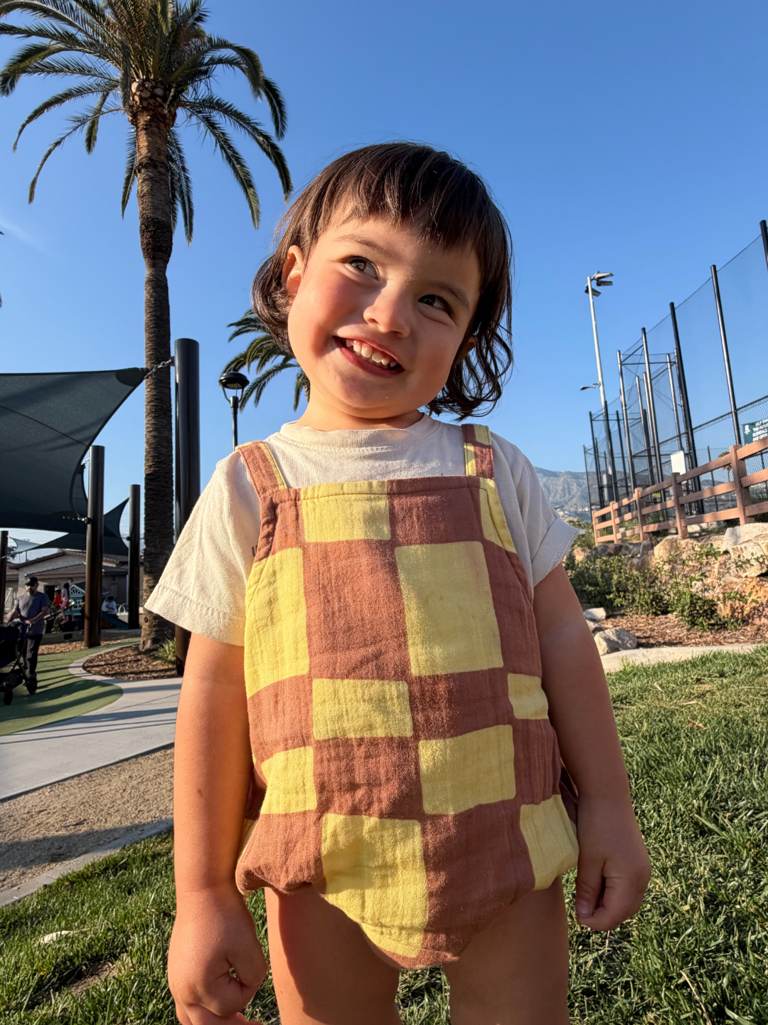 Child wearing a yellow checkered romper standing outdoors with palm trees and a clear sky.