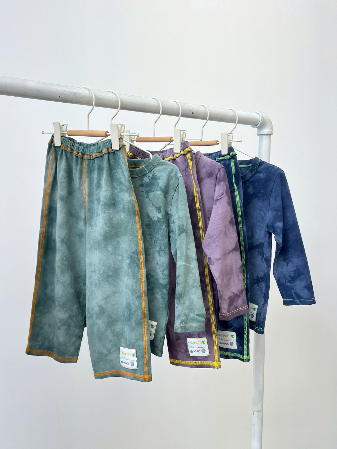 Four COSMO TIE DYE LONG SLEEVE sets for kids in green, purple, navy, and teal with yellow trim hang on a white rack. Each cozy set features a tie-dye longsleeve tee and label, designed in Los Angeles.