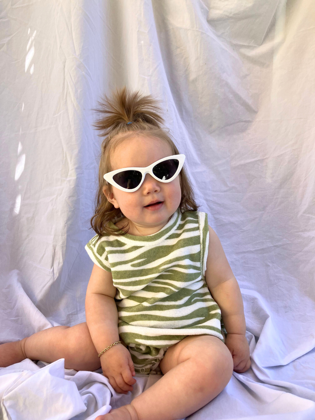 Tiger | A toddler wearing the Terry Bubble Bodysuit in cream terrycloth printed with sage green tiger stripes. She wears white cat eye baby sunglasses, a high ponytail and a gold bracelet, and is seated on a beige cloth backdrop.