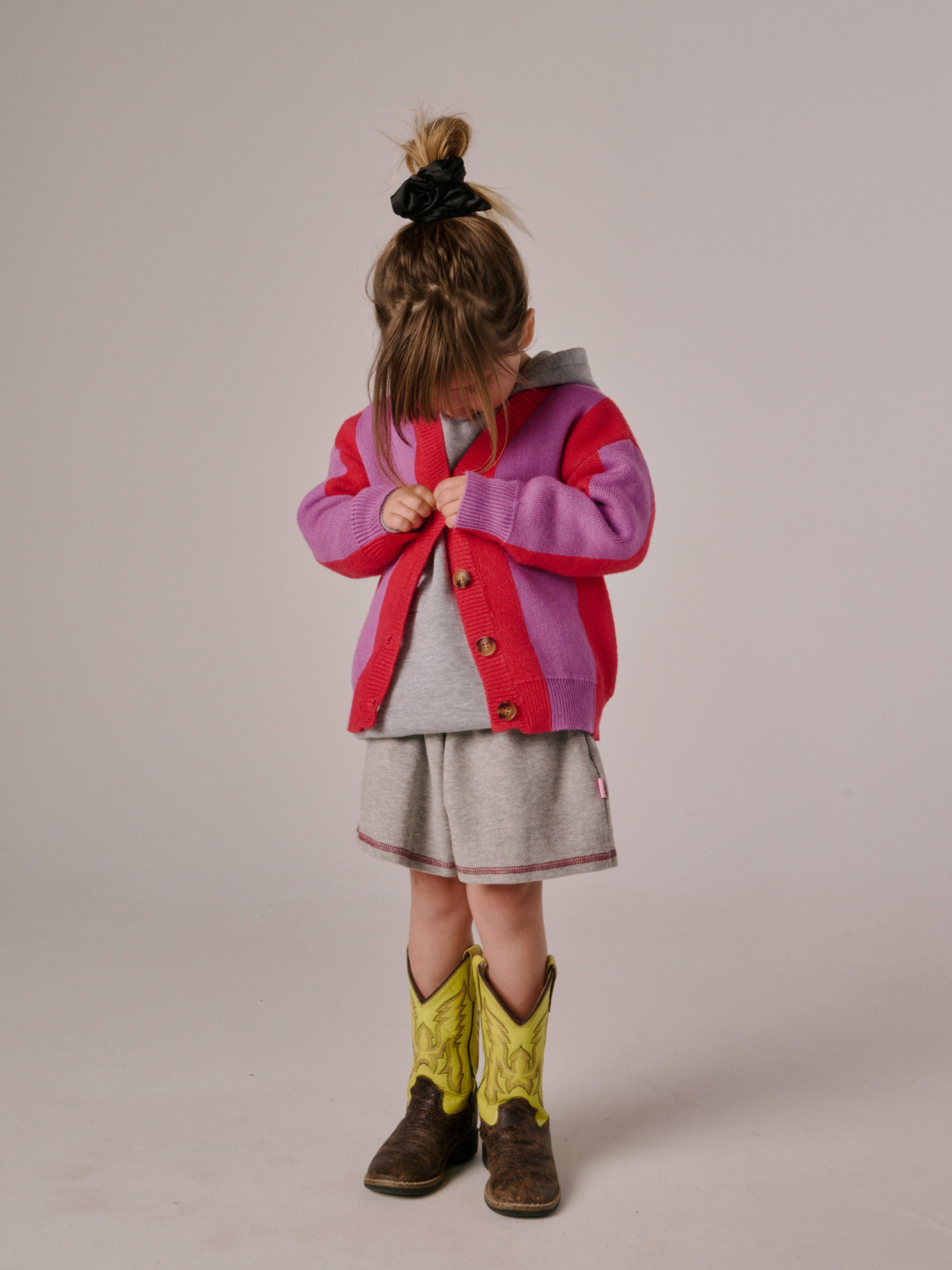 A young child looks down while buttoning the STRIPE CARDIGAN, styled with a grey outfit and yellow cowboy boots, hair in a messy bun.