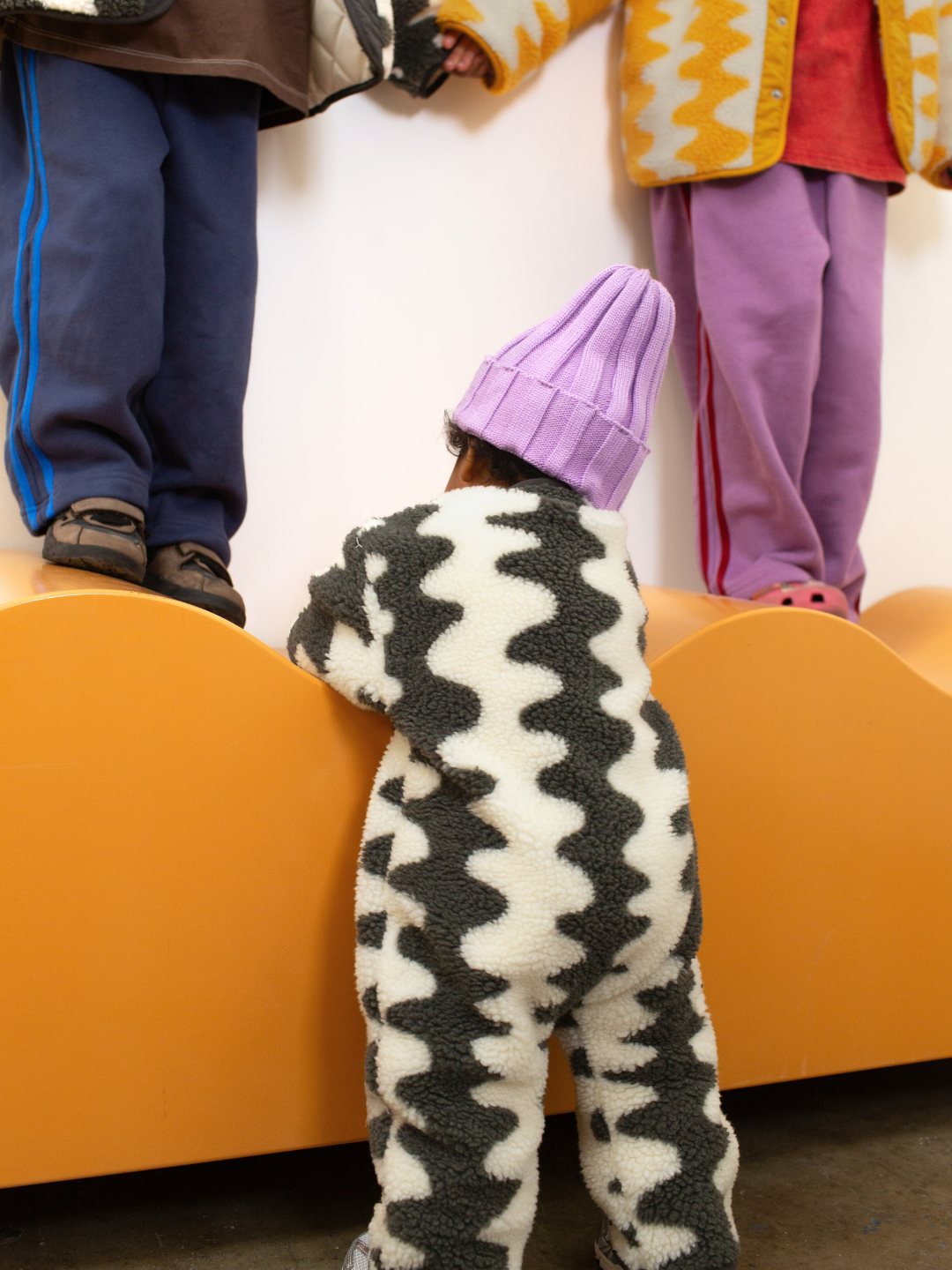Children standing on an orange chair with a white wall in the background, a baby in the wavelength snow suit standing in front. 