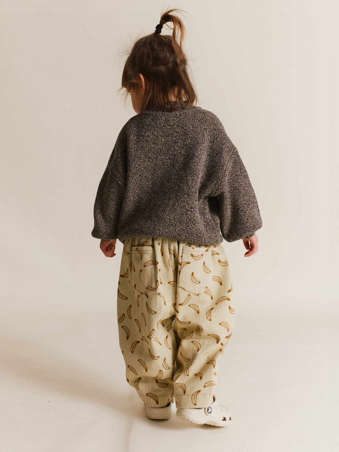 A young child wears the RIAN BANANA TROUSER—oversized balloon leg pants in organic cotton twill with a playful banana print—paired with a loose dark sweater, standing against a plain background, facing away from the camera.