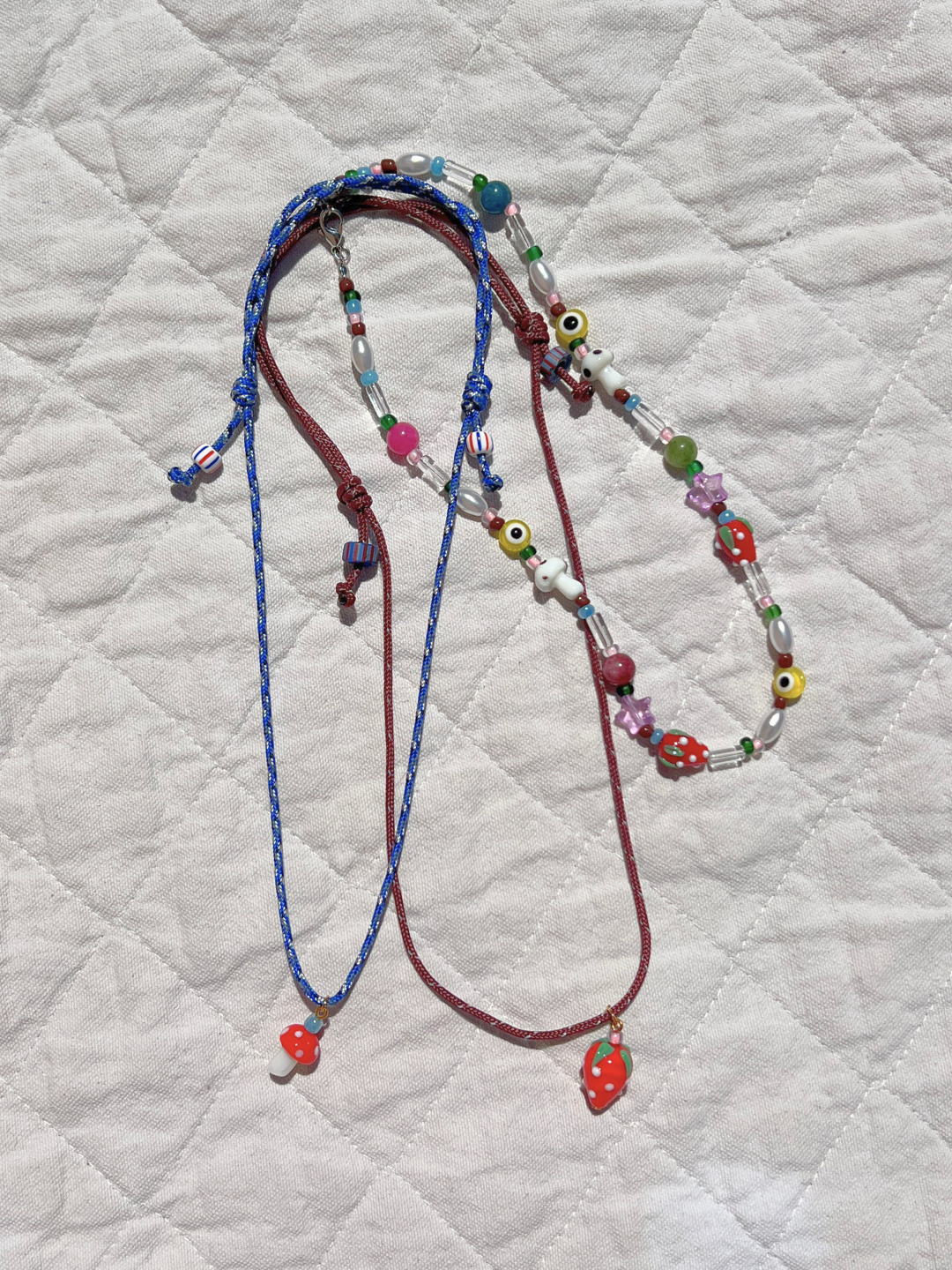 The GAIA NECKLACE features colorful vintage glass beads and mushroom-shaped pendants, displayed on a quilted, light fabric background to showcase this unique, genderless jewelry piece.