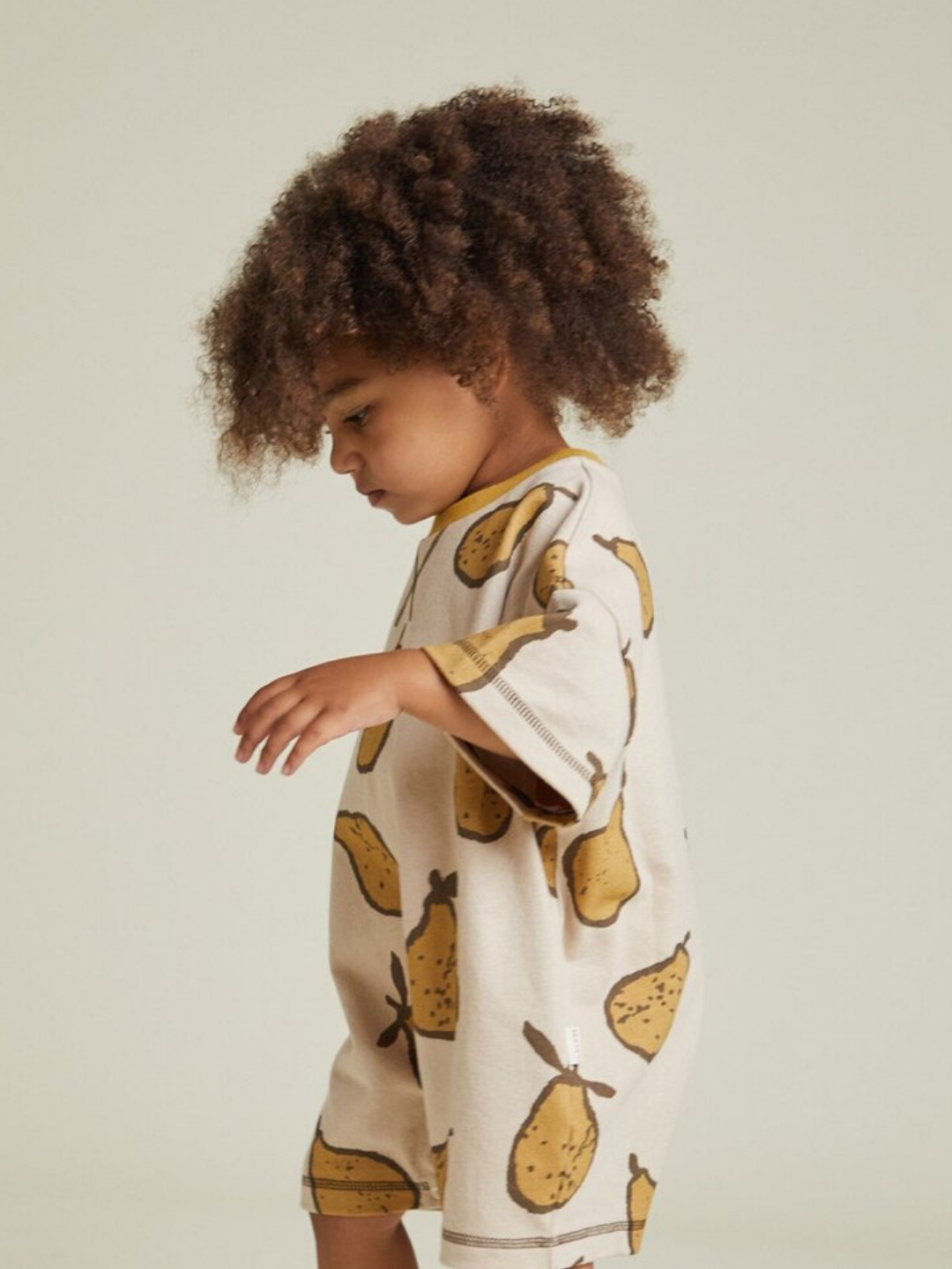A young child with curly hair stands sideways against a plain background, wearing the OPAL ALMOND PEAR ROMPER—a beige romper adorned with yellow pear prints.