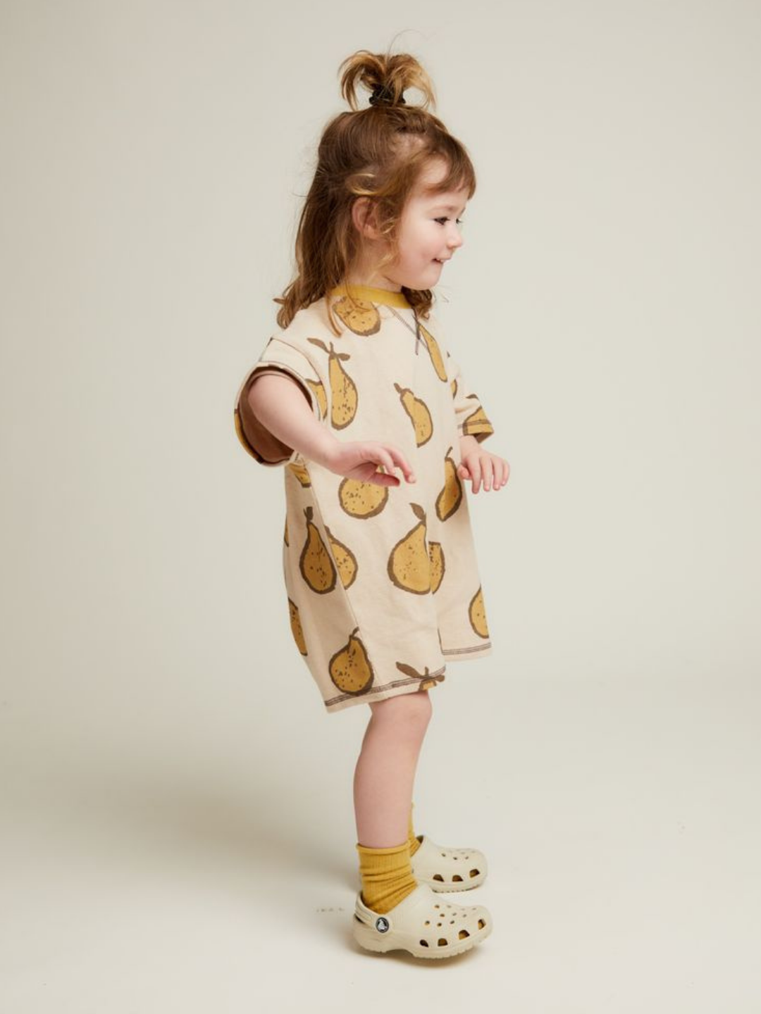 Young child stands sideways wearing the OPAL ALMOND PEAR ROMPER, paired with yellow socks and light clogs, against a plain background.