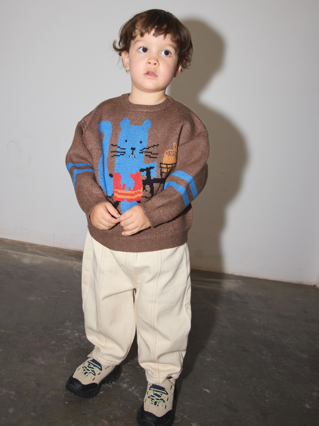 A toddler stands against a plain wall wearing a brown knit sweater with a large blue animal graphic and blue stripe details on the sleeves, paired with cream pants and sneakers.
