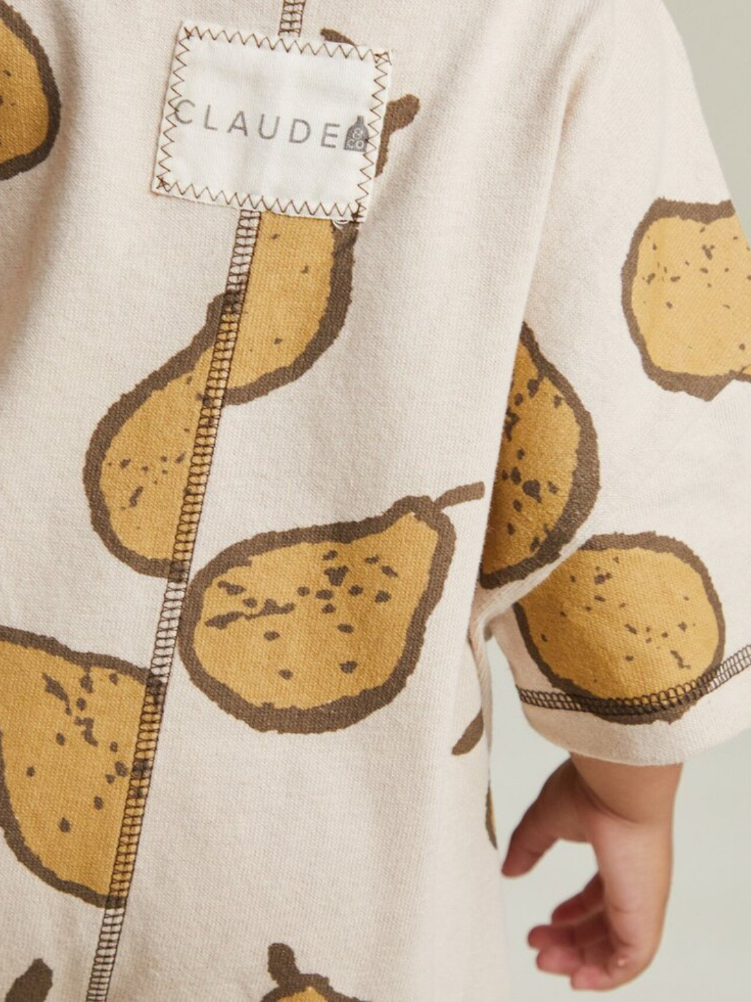 A child wearing the OPAL ALMOND PEAR ROMPER, a beige outfit with brown pear print and a "CLAUDE" label on the back.