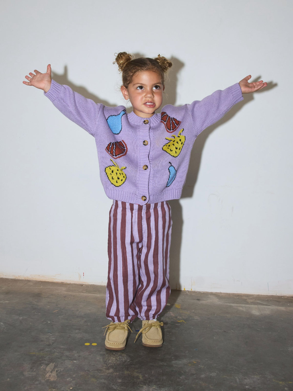Purple Stripe | Child wearing a purple cardigan with a fruit motif and vertically-striped purple and burgundy pants, standing on a grey floor in front of a white wall with her arms raised.