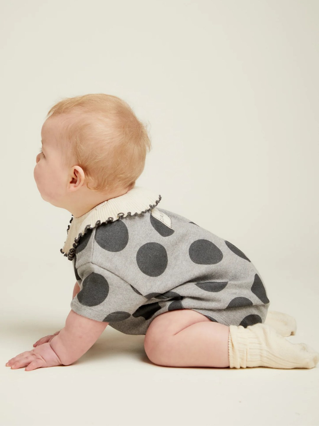 A baby wearing the OPAL MARL SPOT ROMPER, a grey outfit with large black polka dots, and white socks sits on the floor facing left against a plain light background.